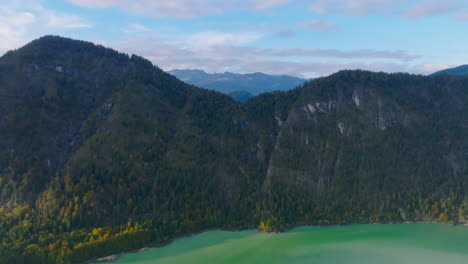 Aerial-view-rising-over-emerald-Stausee-lake-towards-alpine-woodland-Isar-valley-mountain-peak