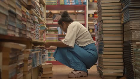 Asian-girl-sitting-and-checking-books-through-bookshelves,-Side-angle-shot