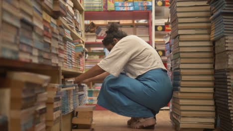 Asian-girl-sitting-and-exploring-books-through-bookshelves,-Side-angle-shot