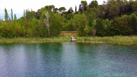 Flying-away-from-a-couple-sitting-in-a-wood-platform-in-front-of-a-lake-with-clear-watter,-between-the-trees-and-house-in-the-background,-in-Banyoles,-Catalonia-,-Spain