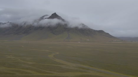 Isolated-house-in-verdant-and-flat-Icelandic-landscape-with-mountains-shrouded-in-low-clouds,-Iceland