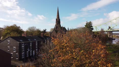 Rising-over-Autumn-trees-to-reveal-small-town-church-with-steeple-alongside-Runcorn-bridge