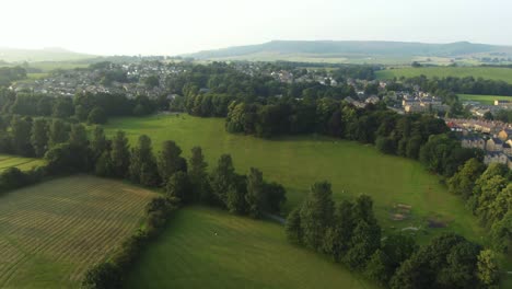 Drone-reveal-of-public-park-at-sunset-surround-by-trees,-hills-and-local-village