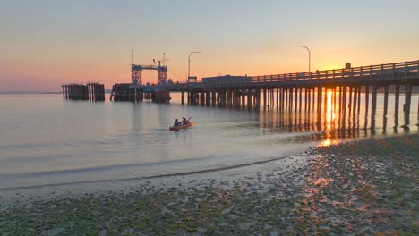 Young-couple-take-off-and-paddle-together-in-sea-kayak-at-sunrise-floating-under-pier-with-sun-glare-reflecting-off-bay-water-near-seattle-washington-aerial-drone-view