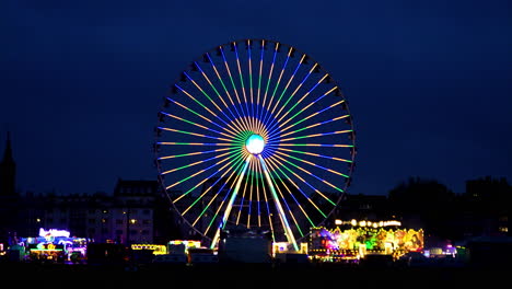 Statische-Handaufnahme-Eines-Leuchtenden-Riesenrads-In-Verschiedenen-Farben-Bei-Einem-Festival-In-Köln-In-Deutschland-Mit-Anderen-Leuchtenden-Fahrgeschäften-Und-Attraktionen-Bei-Nacht