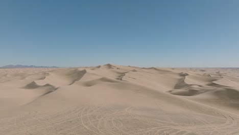 Drone-Shot-Flying-Backwards-Over-Desert-Dunes,-Blue-Sky-Ahead-and-Sandy-Peaks-Below