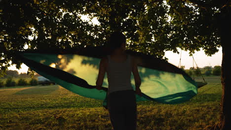 Woman-shakes-out-a-hammock-hanging-between-two-trees-as-the-sun-sets-in-the-background