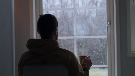 Silhouette-of-man-holding-hot-beverage-and-looking-at-snowfall-through-home-window