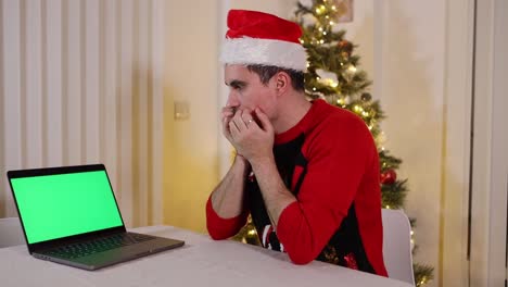 Verängstigter-Junger-Mann-Mit-Weihnachtsmütze,-Der-Am-Tisch-Neben-Dem-Weihnachtsbaum-Sitzt-Und-Auf-Einen-Grünen-Laptop-bildschirm-Schaut