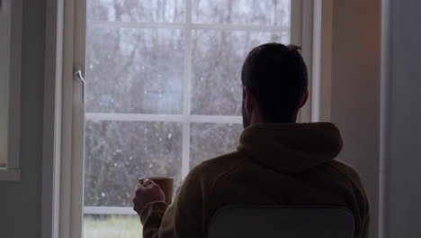 Man-drinking-tea-and-seeing-first-autumn-snow-through-house-window