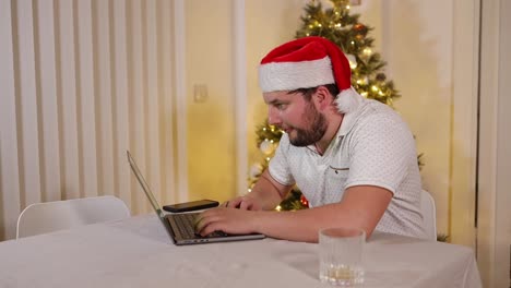 Hardworking-focused-man-with-Santa-hat-sitting-at-table-next-to-Christmas-tree-and-working-on-laptop