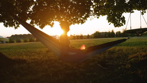 Short-haired-woman-sits-up-in-a-hammock,-looking-out-over-a-sunset-casting-a-large-lens-flare