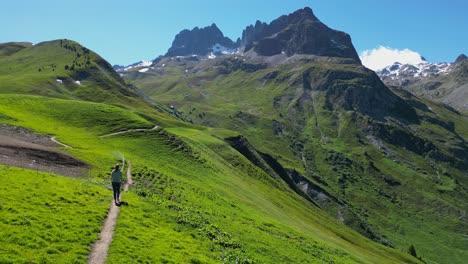 Young-Adult-Woman-hikes-green-valley-to-mountains-in-French-Alps---Aerial-Follow