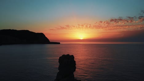 Schöner-Orangefarbener-Sonnenuntergangshimmel-Am-Horizont-Mit-Antennenwagen-Zurück-über-Silhouettierten-Felsen-Vor-Dem-Strand-Von-Cala-Benirras-Auf-Ibiza