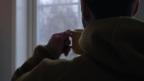 Close-up-over-shoulder-shot-of-man-with-warm-drink-looking-snow-falling-in-winter