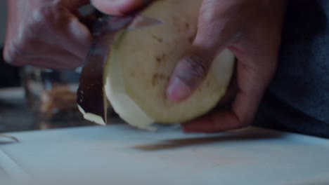 Young-black-man-peeling-ripe-half-eggplant-on-plastic-white-board-in-kitchen-at-night