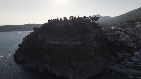 Aerial-Cinematic-Silhouette-View-Of-Hillside-With-Parga-Castle,-Slow-Parallax-To-Reveal-Sun-Shining-On-Island