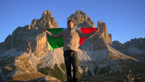 Footage-filmed-at-Rifugio-Auronzo,-Tri-Cine-up-the-mountains-in-Italian-Dolomites-with-a-man-moving-and-holding-an-Italian-flag-and-with-beautifull-mountains-in-the-backgorund,-filmed-at-sunrise-in-4k