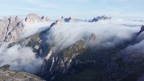 Low-clouds-at-sunrise-flowing-over-jagged-landscape-of-Italian-Dolomites,-aerial