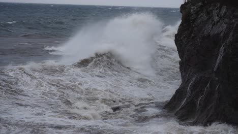 Rough-waves-crash-against-rock-coastal-cliff-on-a-gloomy-day,-slow-motion