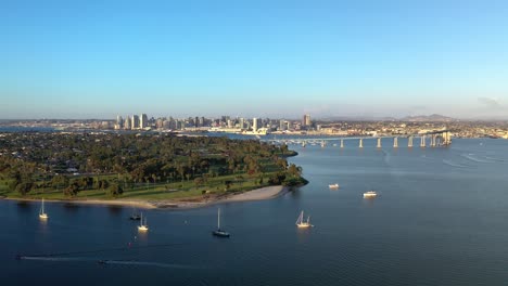 Still-Shot-of-Coronado-Bridge-and-San-Diego-Skyline-During-Golden-Hour-with-Boats-Sailing-by-on-Ocean-with-Trees-and-Park-on-the-Left