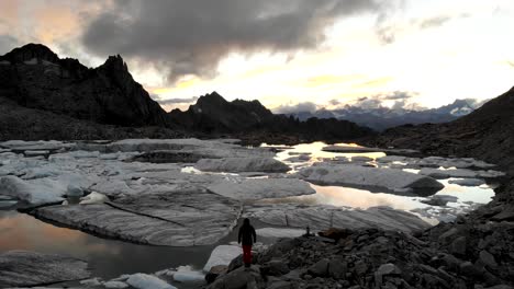 Aerial-view-of-a-hiker-looking-a-glacier-lake-full-of-melted-icebergs-in-remote-parts-of-the-Swiss-Alps-with-sunset-glow-reflecting-on-the-water