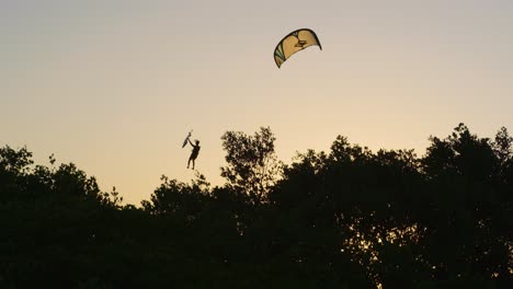 Silhouette-Kitesurfer-jumping-in-sky-during-golden-sunset-behind-forest-trees---static-wide-shot-in-slow-motion