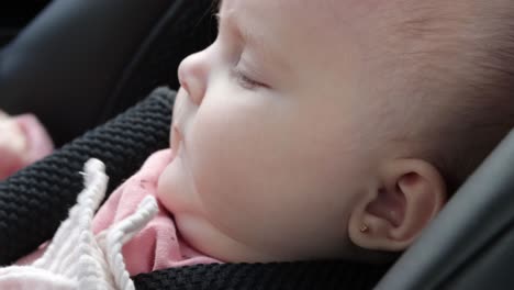 Tired-baby-girl-in-car-seat-sucks-fingers,-rubs-her-eyes,-looking-at-camera
