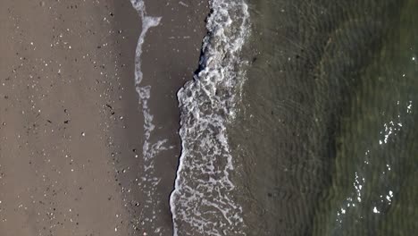 A-top-down-view-over-an-empty-beach-as-the-waves-gently-crash-onto-the-shore-in-slow-motion