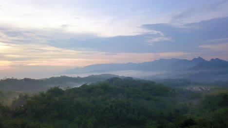 Enthüllen-Sie-Drohnenaufnahmen-Von-Wäldern,-Die-Von-Nebel-Mit-Sonnenaufgangshimmel-Im-Hintergrund-Umhüllt-Sind---Tropische-Landschaft-Indonesiens-An-Einem-Nebligen-Morgen