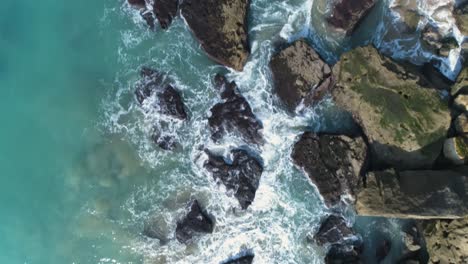 Aerial-view-of-a-beautiful-turquoise-blue-sea-waves-crashing-on-rocks-on-a-sunny-day