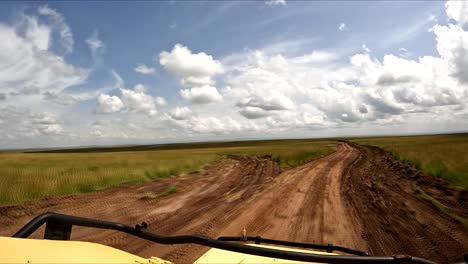 Safari-drive-in-Maasai-Mara-plains-through-dirt-road-with-blue-sky-and-big-clouds