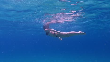 Underwater-scene-beneath-seawater-surface-of-adult-woman-swimming-with-black-bikini-in-crystal-clear-deep-blue-ocean-water-with-diving-mask