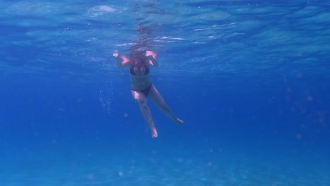Underwater-scene-beneath-seawater-surface-of-adult-woman-with-black-bikini-floating-and-swimming-in-crystalline-deep-blue-ocean-water