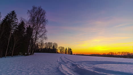 Goldener-Orangefarbener-Sonnenuntergangshimmel-Im-Zeitraffer-über-Schneebedecktes-Feld,-Gesäumt-Von-Silhouetten-Von-Bäumen