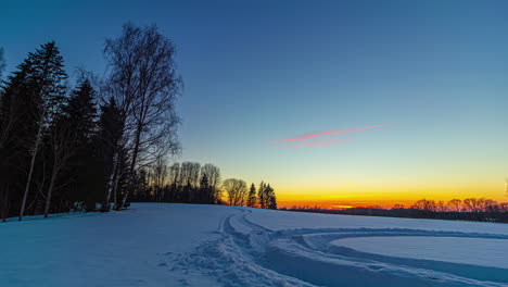 Filmischer-Sonnenuntergang-Im-Zeitraffer-Am-Horizont-Durch-Schneebedeckte-Landschaft-Mit-Silhouetten-Von-Tannen-Und-Reifenabdrücken-Im-Vordergrund