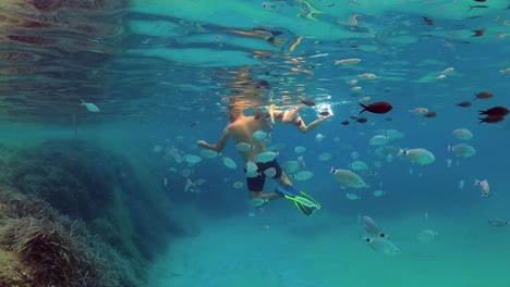 Underwater-slow-motion-scenic-view-of-father-and-little-son-swimming-in-blue-tropical-sea-water-surrounded-by-school-of-fish