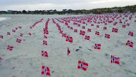 Aerial-view-flying-over-multiple-Norway-flags-on-sandy-beach-coastline-blowing-in-wind