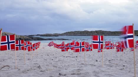 Low-panning-shot-across-multiple-rows-of-Norway-flags-on-sandy-coastal-beach-blowing-in-breeze