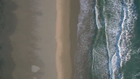 Aerial-drone-shot-looking-down,-symmetrically-aligned-with-beach-shore-and-sea-waves-crashing