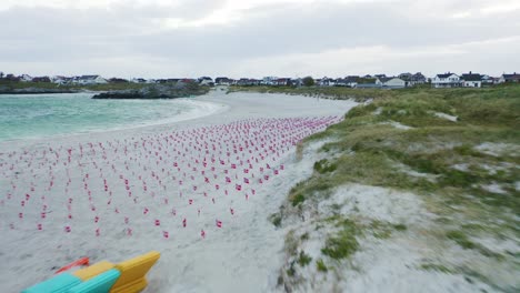 Aerial-view-rising-pull-back-reveal-across-Norway-coastline-passing-many-rows-of-Norway-flags-blowing-on-sandy-beach