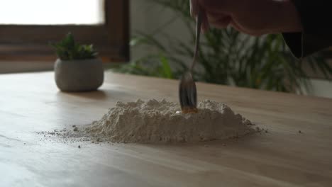 Person-preparing-mix-blending-flour-and-egg-yolk-on-wooden-kitchen-table-surface-making-fresh-pasta-dough