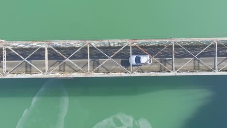 Two-cars-driving-through-narrow-metal-baltimore-type-bridge-on-lake,-overhead-static-aerial