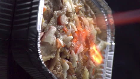 Dutch-angle-view-of-chicken-mentai-mixed-with-colorful-vegetables-cooking-with-hot-burning-torch-flame-of-fire-sizzling-surface-of-food-in-silver-foil-take-out-pan,-static-close-up