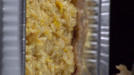 Dutch-angle-vertical-view-of-cooked-slices-of-meat-being-added-and-layered-above-yellow-rice-surface-with-tongs-in-silver-foil-take-out-pan,-static-extreme-close-up-detail