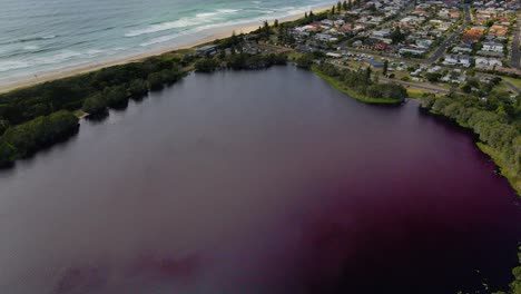Beautiful-red-Lake-Ainsworth-by-the-waves-and-town-of-Lennox-Head,-Australia--aerial