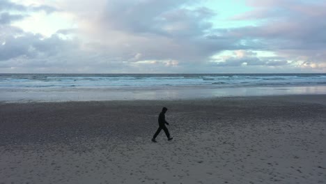 A-man-wearing-black-only-walks-by-himself-on-a-beach-with-a-dreamy-sky,-aerial