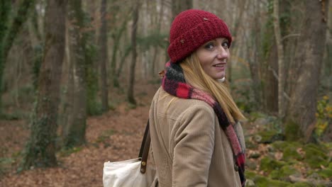 Woman-in-red-hat-turns-to-smile-at-camera-while-walking-in-autumn-forest,-Slowmo