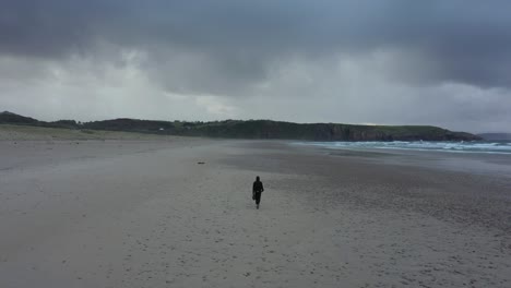 Back-view-of-man-walking-alone-on-empty-deserted-beach-on-winter-cloudy-day