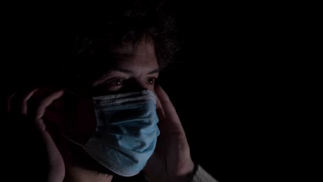 Young-bearded-male-putting-on-corona-virus-surgical-mask-in-darkness
